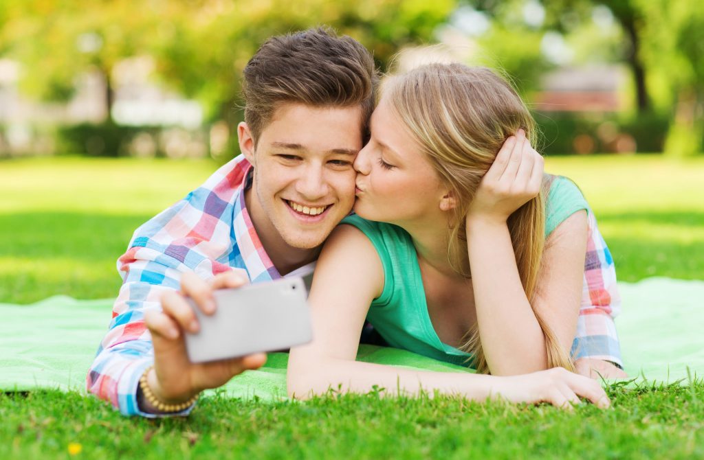 couple laying on a blanket in the grass taking a selfie. The woman is kissing the man on the cheek